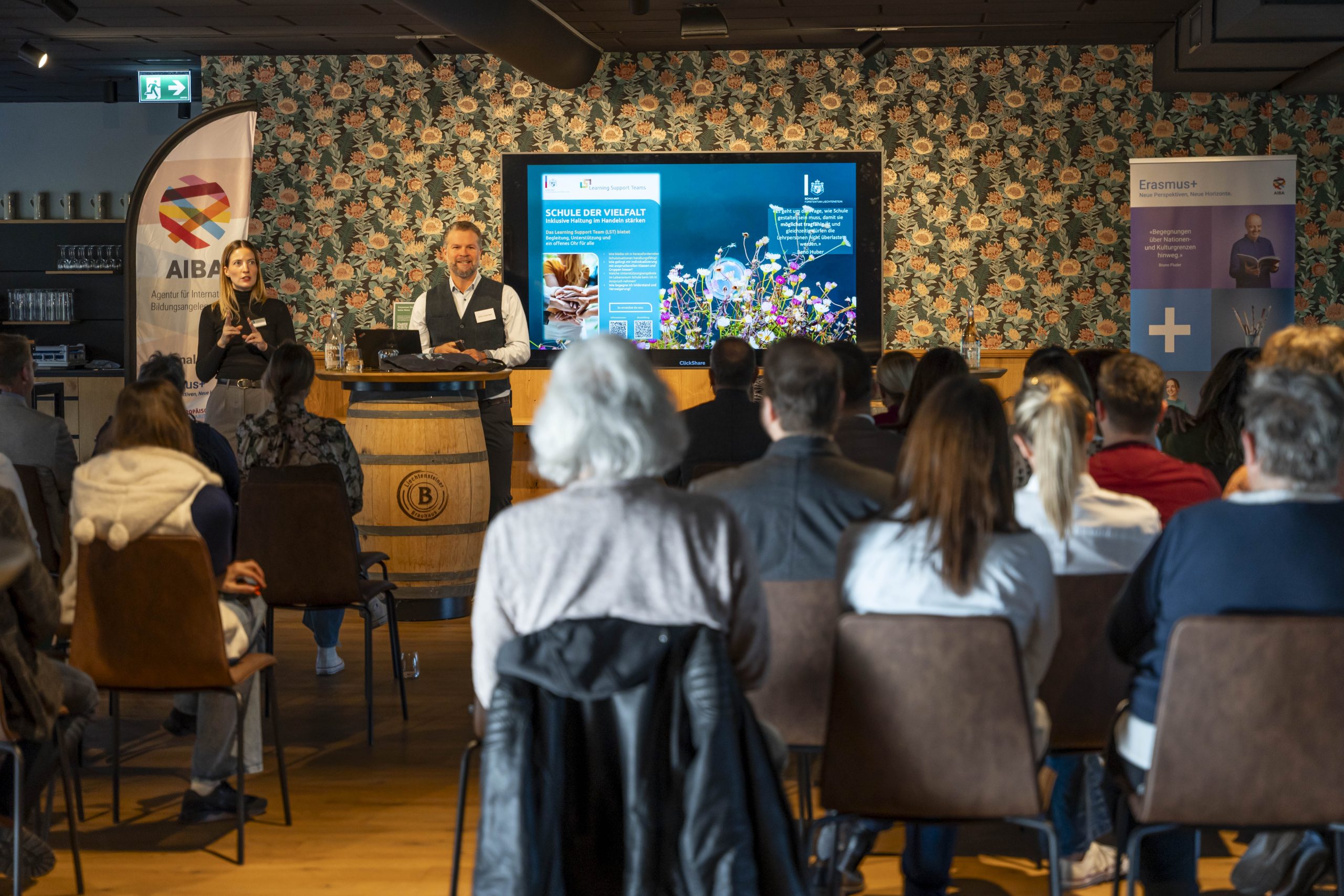 Präsentation vor Publikum mit grossem Bildschirm und Infobannern in modernem Raum.