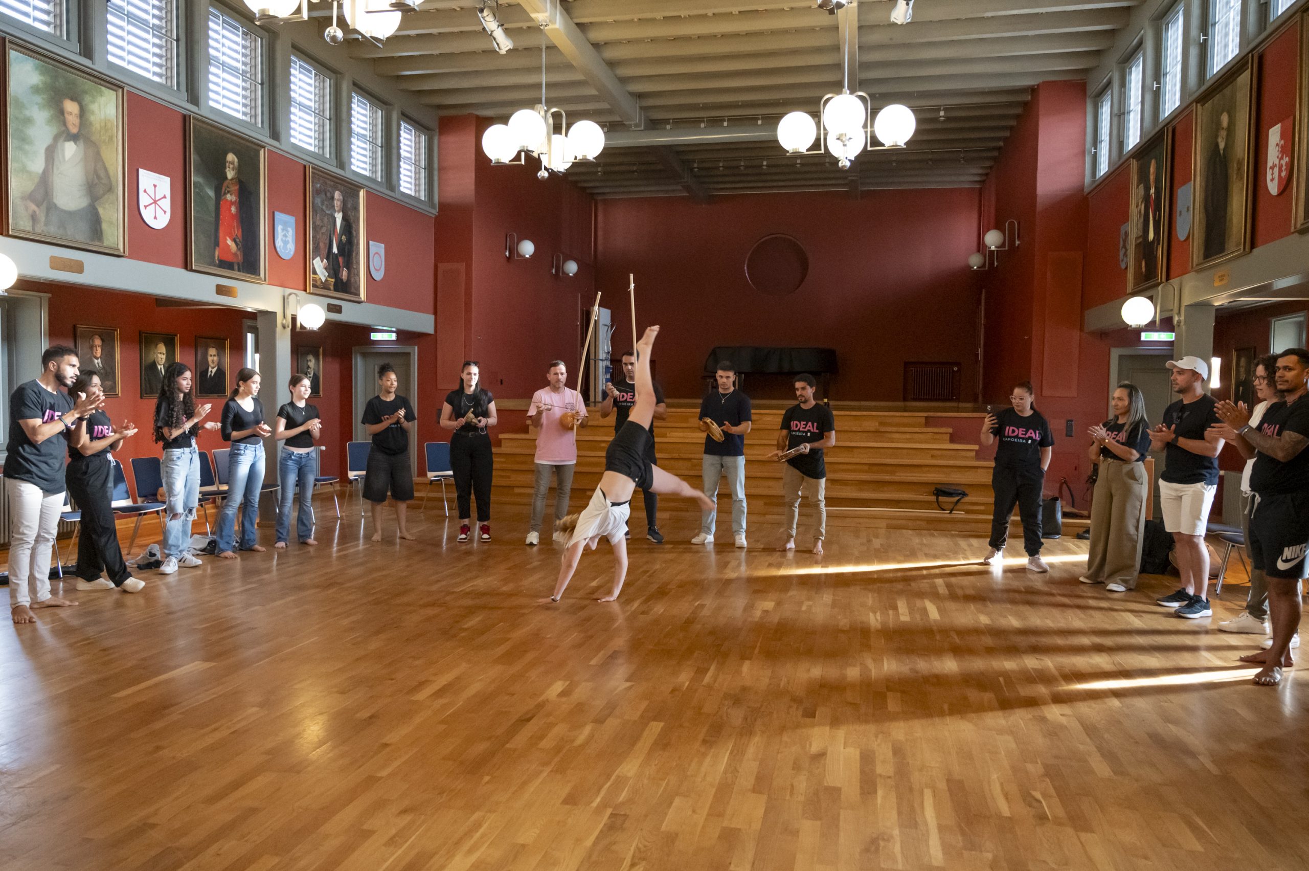Gruppe von Menschen steht im Kreis im Rathaussaal Vaduz, während eine Person in der Mitte eine Capoeira-Bewegung ausführt