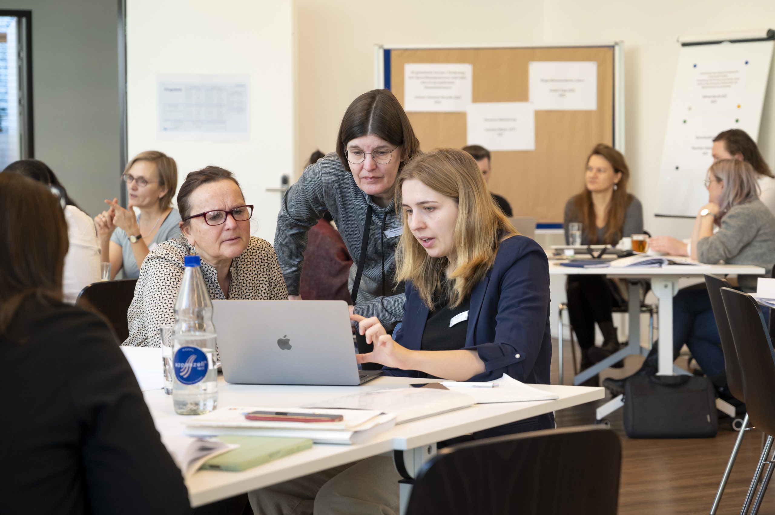 Drei Personen arbeiten gemeinsam an einem Laptop, im Hintergrund sind weitere Personen zu sehen