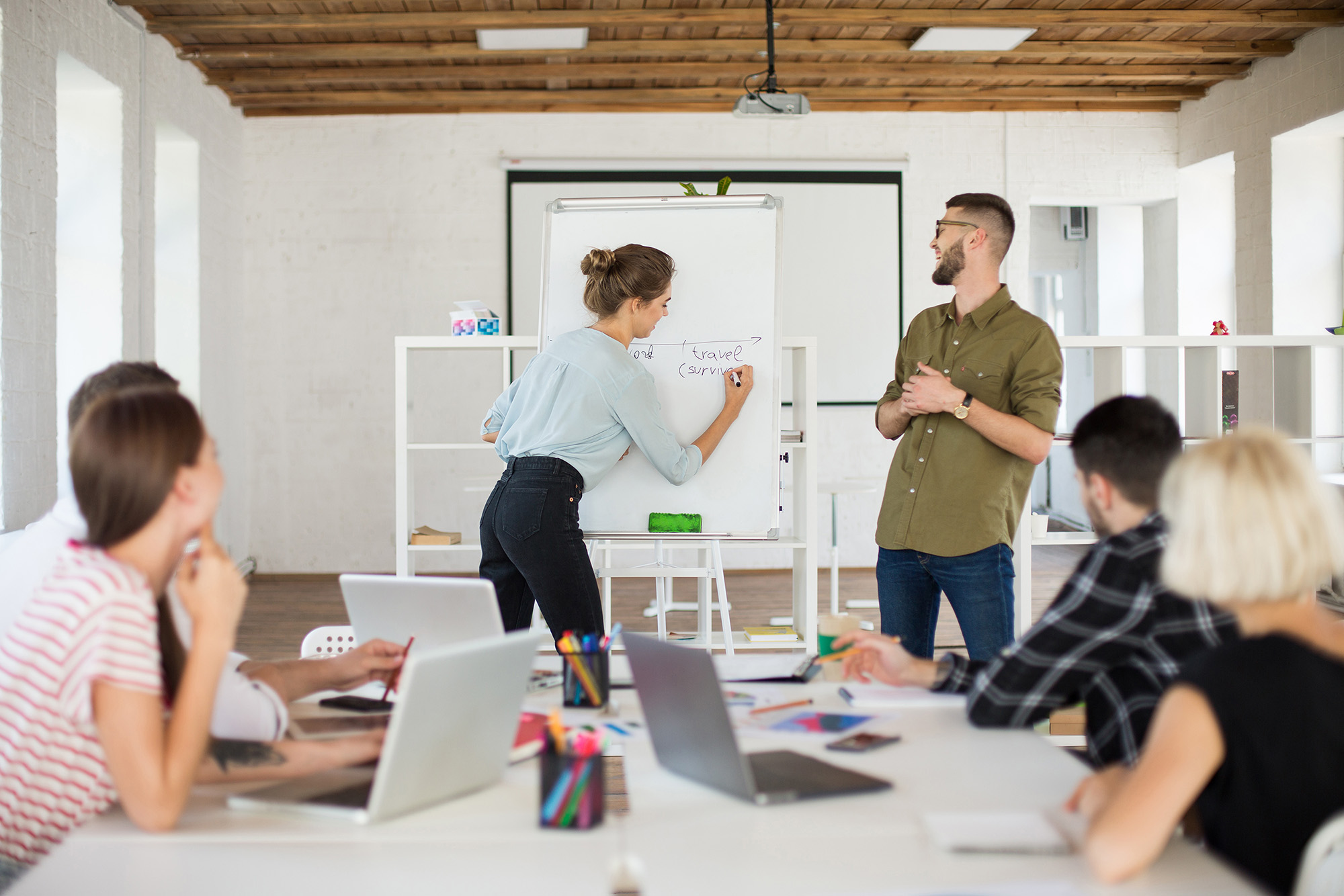 Eine Frau schreibt auf einem Whiteboard, ein Mann steht neben der Tafel und weitere Personen sitzen an einem Tisch und schauen zu