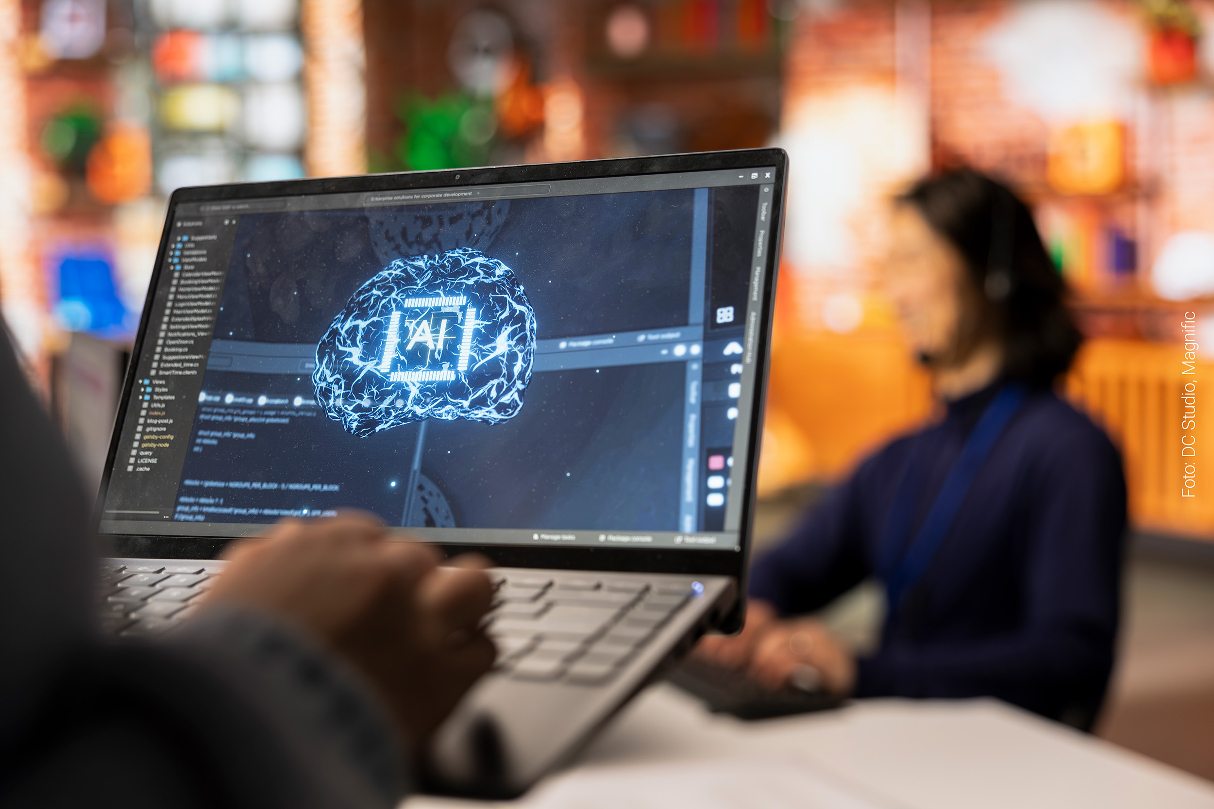 Person arbeitet an Laptop, auf dem die Buchstaben "AI" leuchten, im Hintergrund sitzt eine Person an einem Laptop