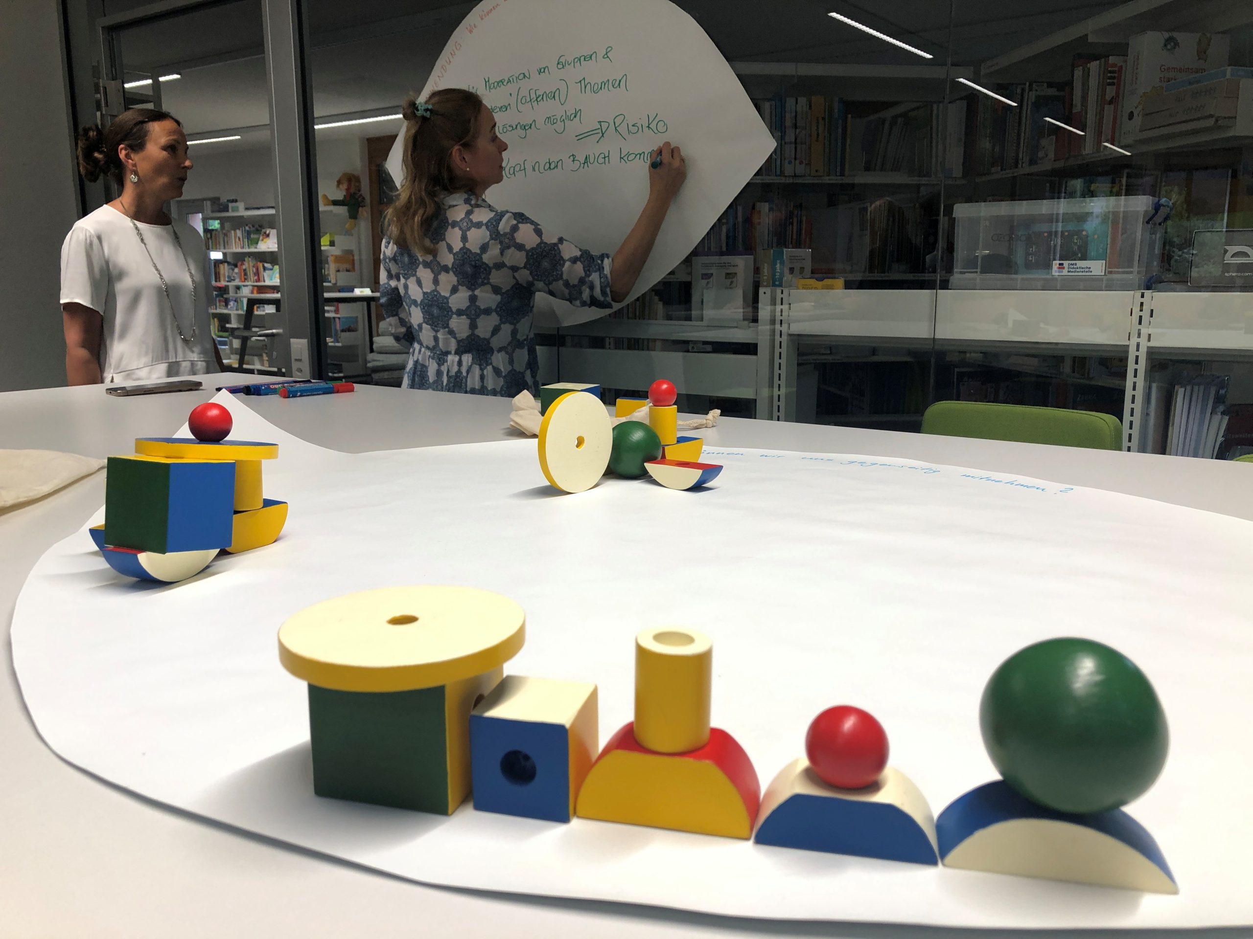 Zwei Personen arbeiten in einem Besprechungsraum. Auf einem grossen Tisch liegt ein rundes Blatt, auf dem farbige Holzbausteine angeordnet sind.