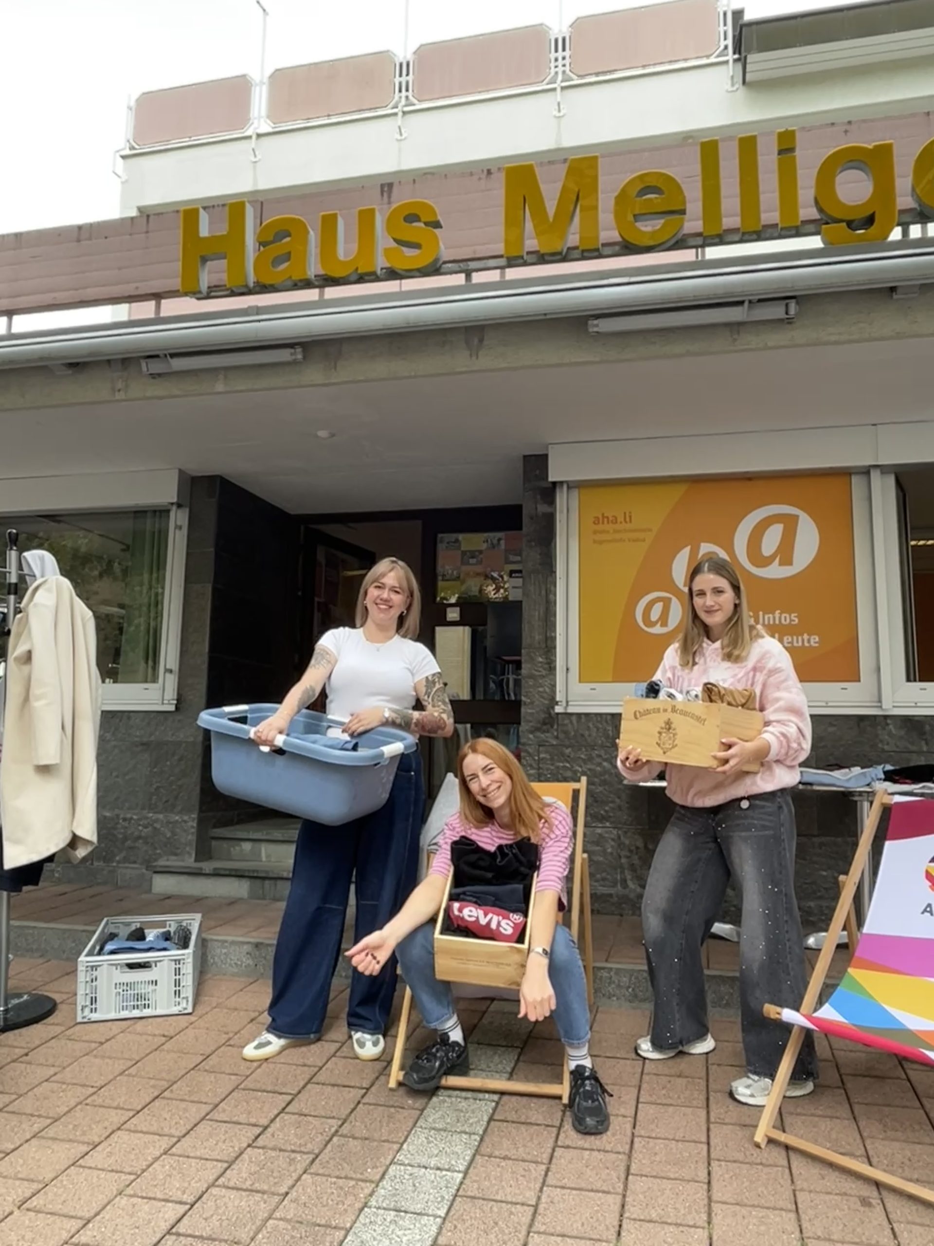 Drei Personen sitzen bzw. stehen vor dem aha mit Kleiderkisten in den Händen und sind bereit für den Parking Day 2026